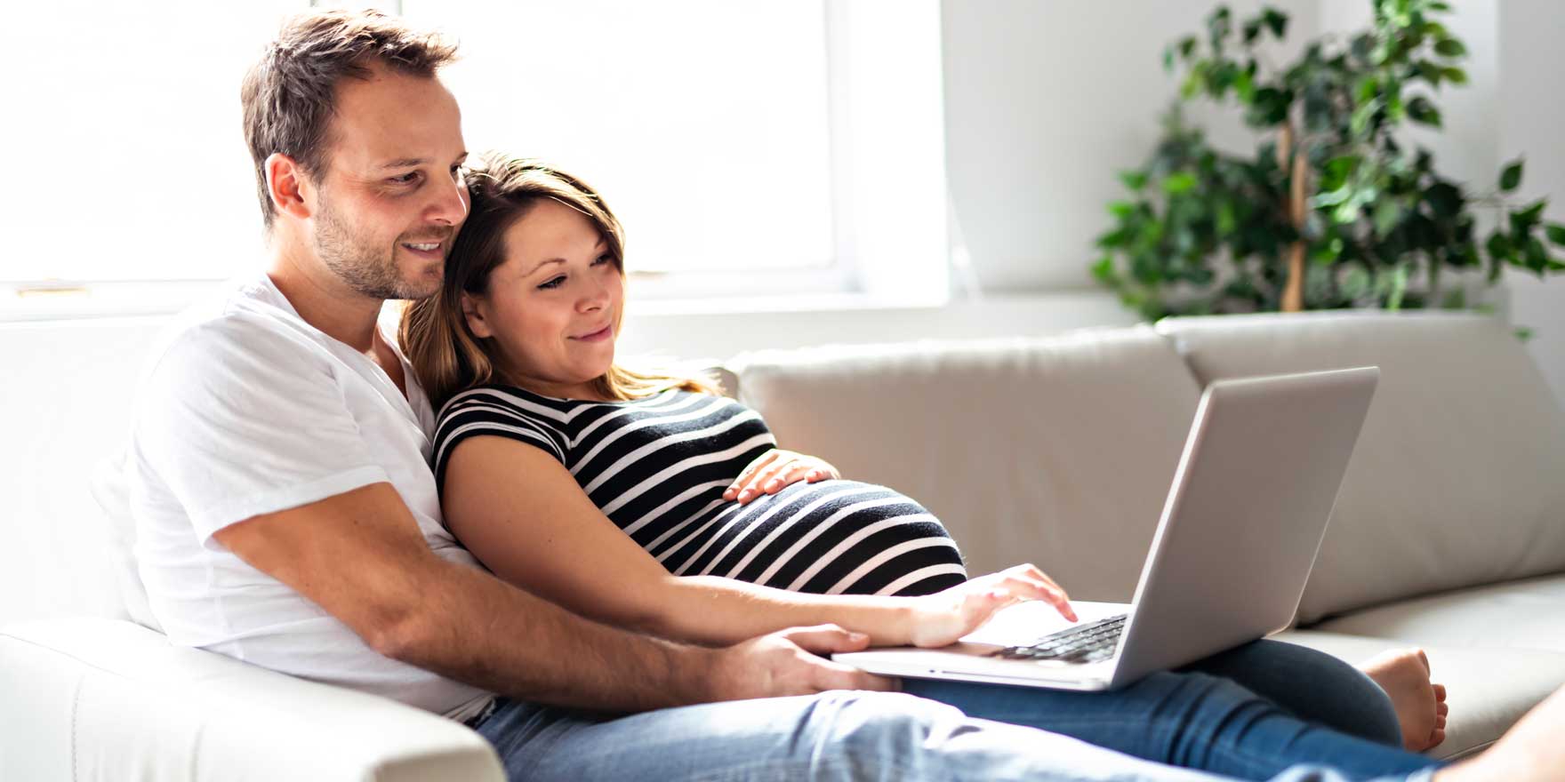 Pregnant mom and dad sitting on a sofa while looking at a laptop screen together