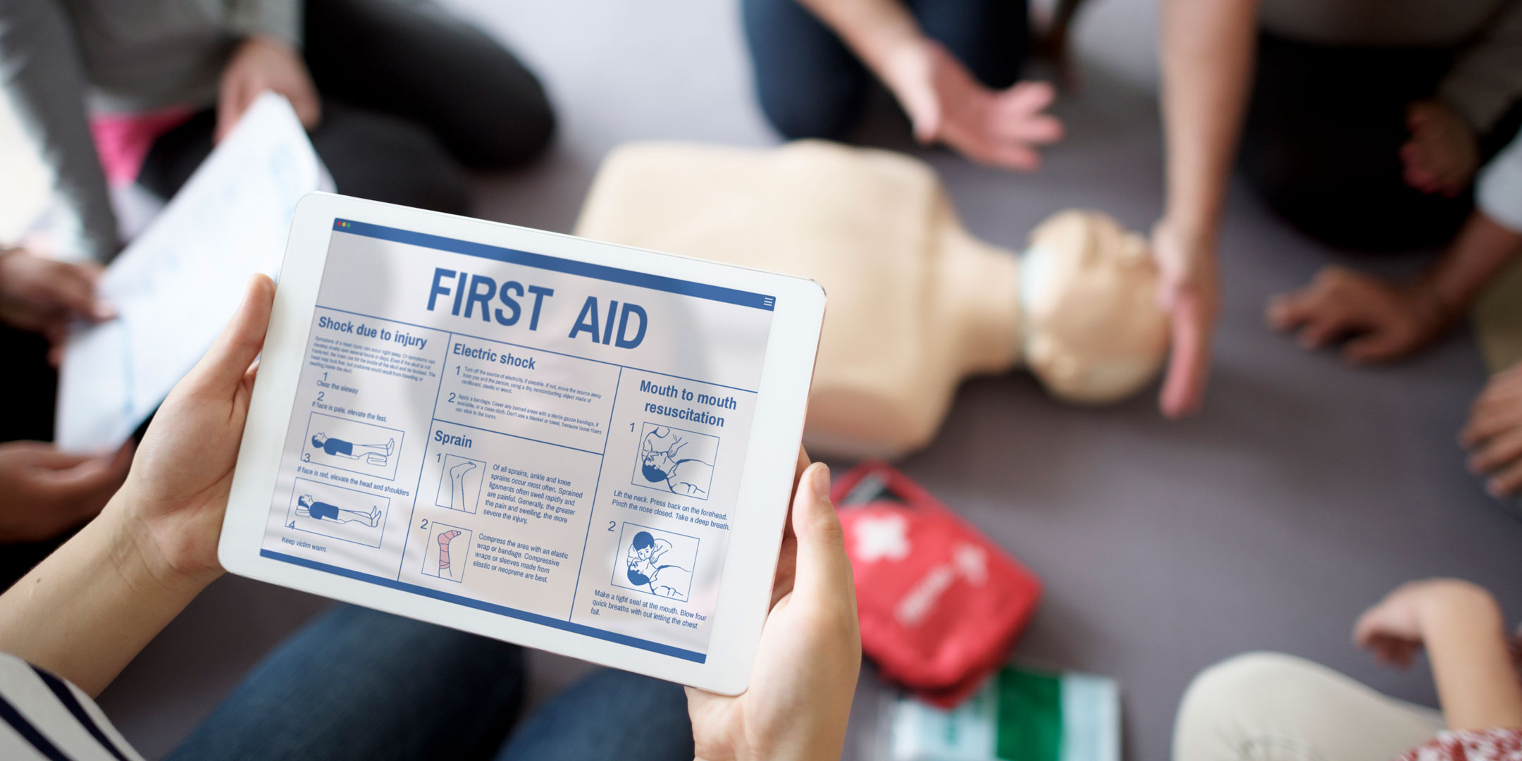 Close-up for a person looking at checklist for First Aid while an instructor provides a how-to lesson on the floor with a manikin and a first aid kit