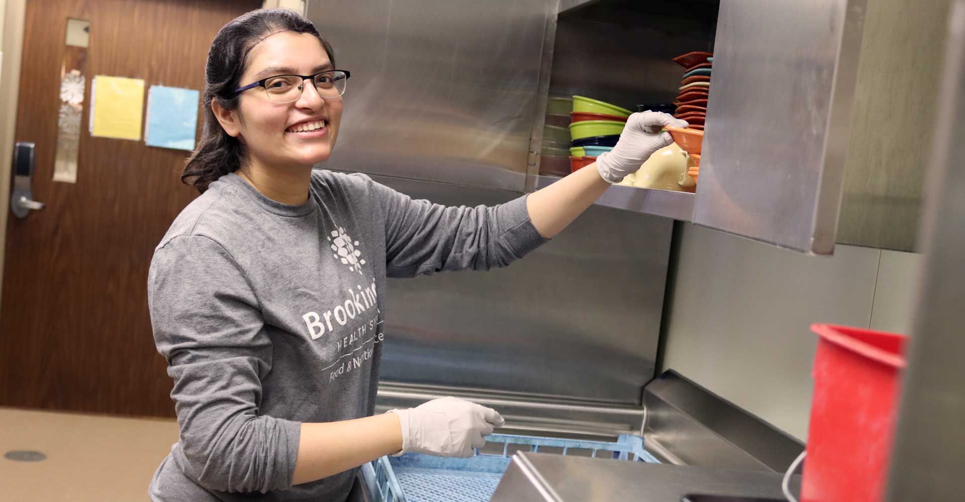 Janhavi, food service worker at The Neighborhoods, putting away dishes in the kitchen and smiling at the camera