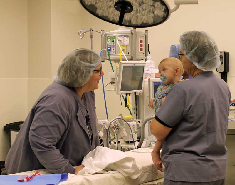 A toddler being held by an OR nurse inside an operating room while another operating room nurse gives him a friendly smile
