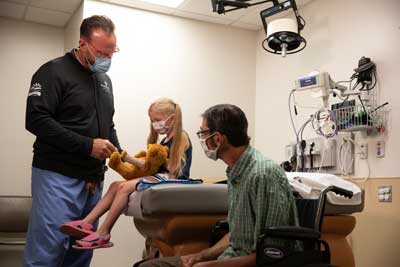 Medical provider showing a young girl how she'll get a bandage by demonstrating on a bear while dad in a wheel chair looks on